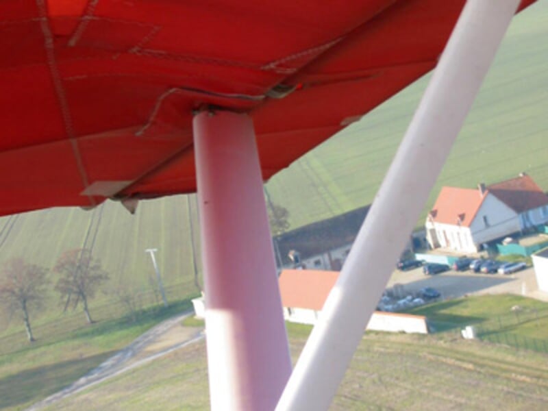 Vue partielle d'un ULM rouge en vol au-dessus d'un paysage rural avec des maisons et des champs verts.